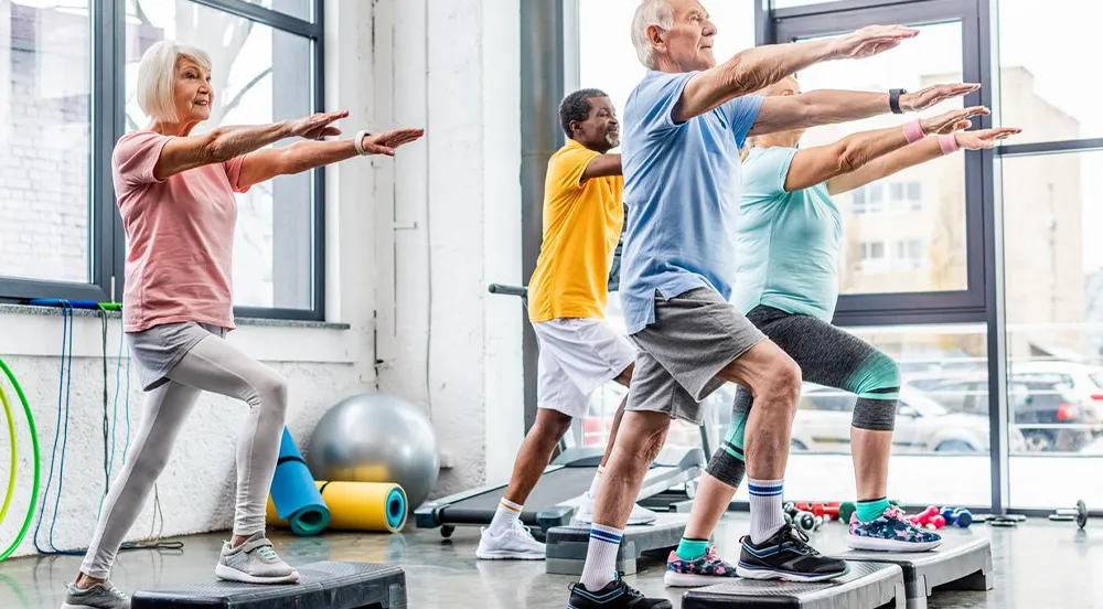 Personnes âgées faisant des exercices de step dans une salle lumineuse, portant des vêtements colorés. Ambiance dynamique et motivée.