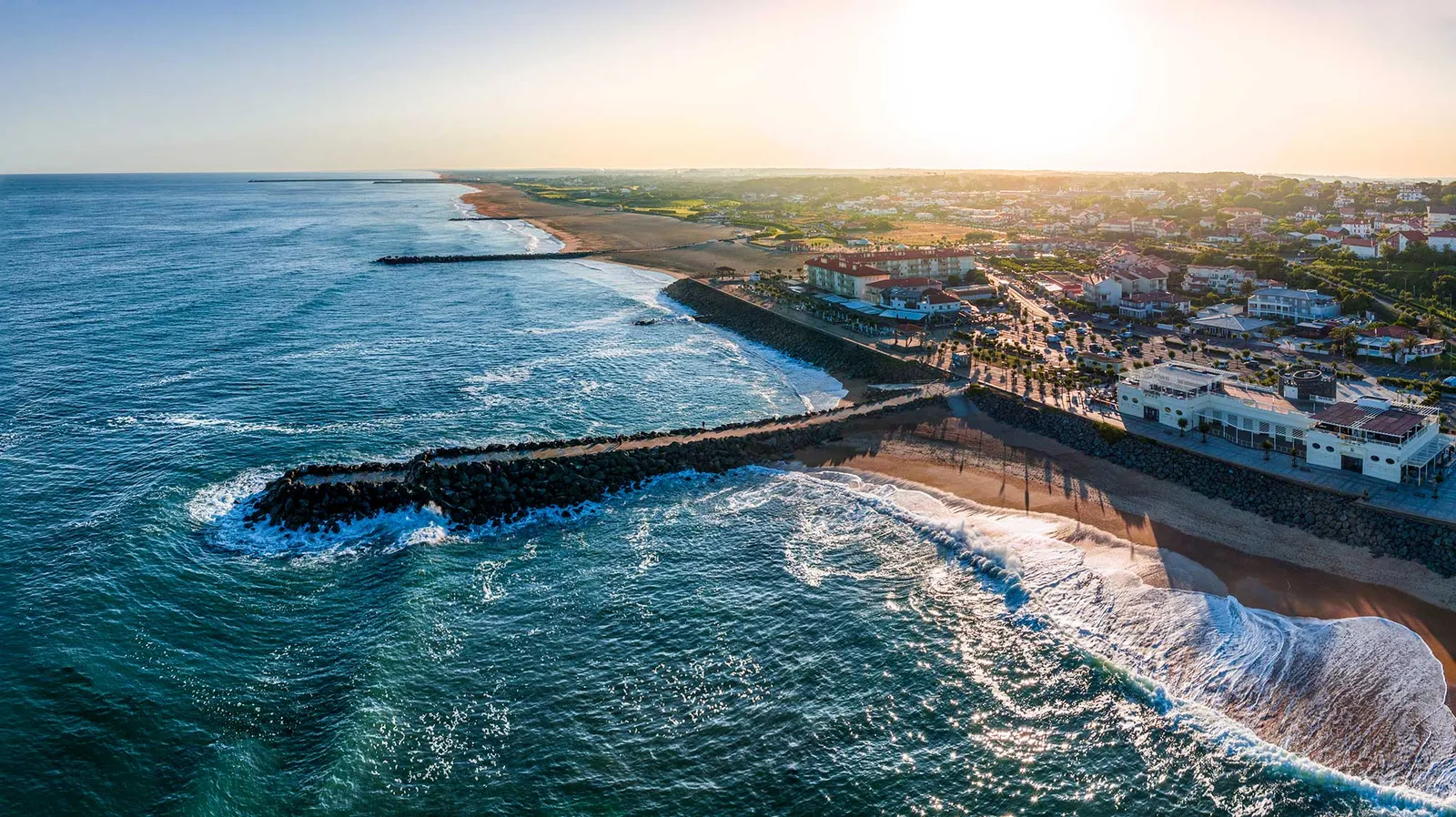 Vue aérienne d'une plage et d'une ville côtière au coucher du soleil. Les vagues déferlent sur la jetée rocheuse. Ciel clair et lumineux.