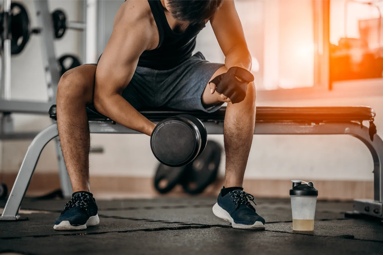 Personne assise sur un banc de gym, soulevant un haltère. Gant noir, short gris. Gourde au sol. Ambiance sportive avec éclairage chaud.