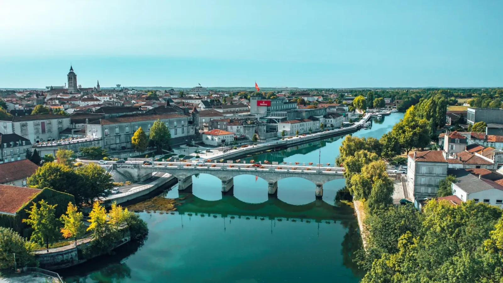 Vue aérienne d'une ville avec une rivière et un pont. Bâtiments anciens, arbres verdoyants, ciel bleu. Ambiance paisible et estivale.