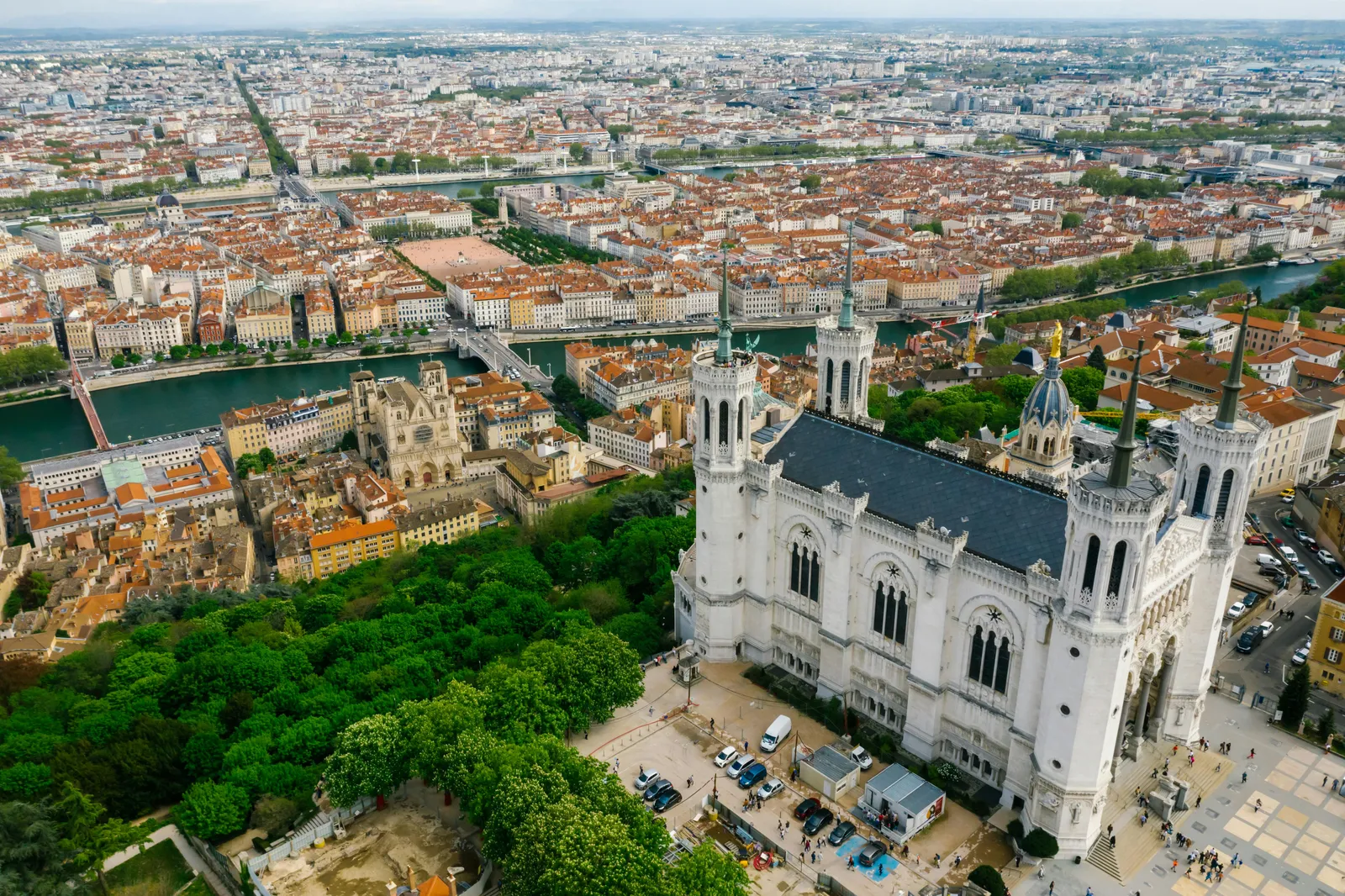 Vue aérienne de Lyon, montrant la basilique Fourvière blanche au milieu, entourée de verdure et de bâtiments rouges. Ciel nuageux.